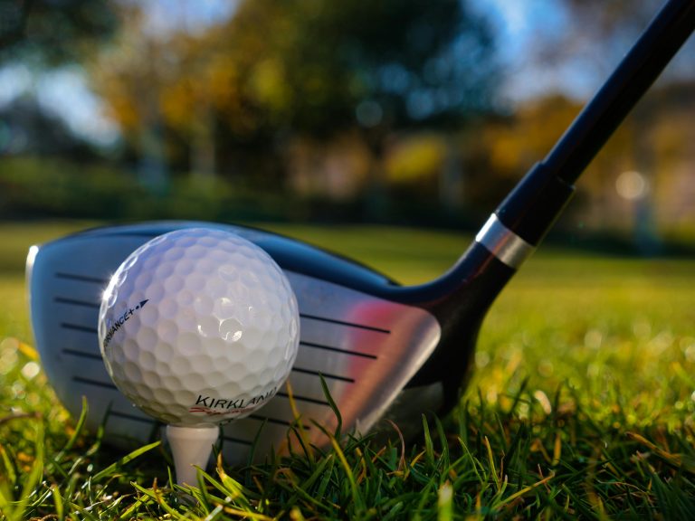 Close-up of a golf ball on a tee with a driver clubface positioned square behind it on grass